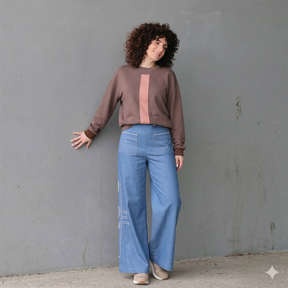 Een jonge vrouw met krullend haar leunt lachend in atelier van CLAU-D in Arnhem. Ze draagt een oversized tweekleurige basic sweater met een zwart labyrint-motief en een bijpassende blauw bewerkte basic jeans broek uit de collectie van Claudia Träumer.
